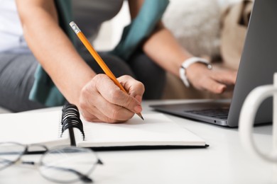 Photo of Woman learning online using laptop and taking notes at white table indoors, closeup. Self-study