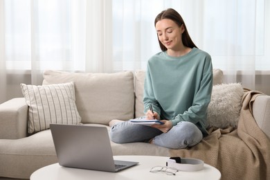 Photo of Woman taking notes during online lesson on sofa at table indoors. Self-study