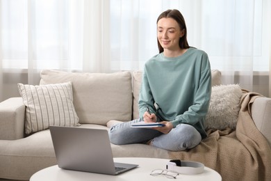 Photo of Smiling woman taking notes during online lesson on sofa indoors. Self-study