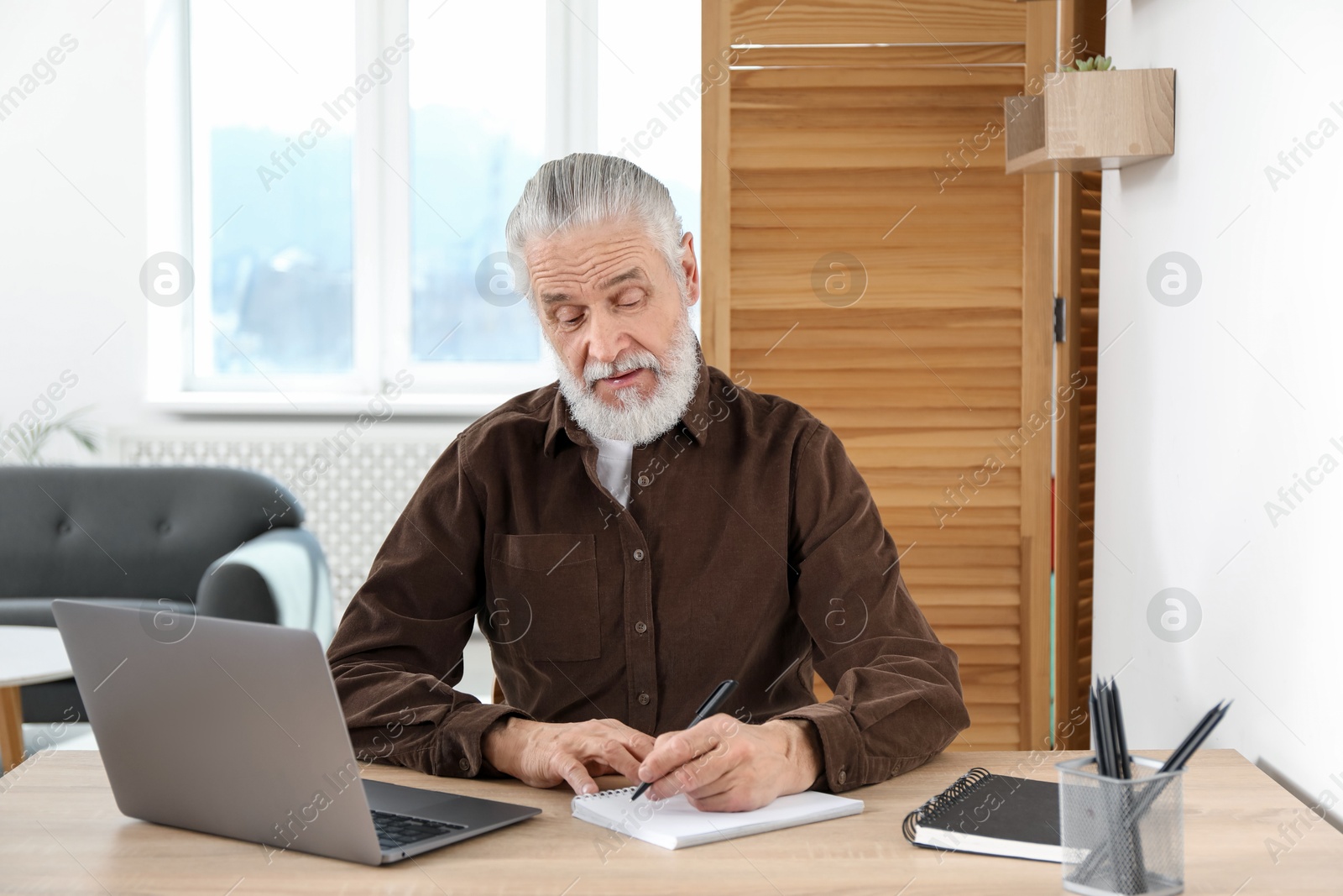 Photo of Senior man taking notes while learning online at desk indoors. Self-study