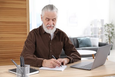 Photo of Senior man taking notes while learning online at desk indoors. Self-study