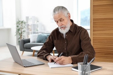 Photo of Senior man taking notes while learning online at desk indoors. Self-study