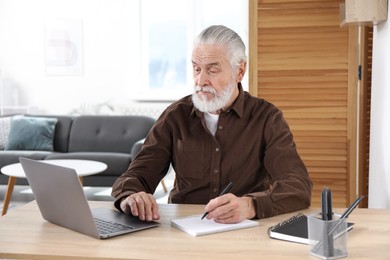 Photo of Senior man taking notes while learning online at desk indoors. Self-study