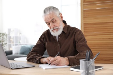 Photo of Senior man taking notes while learning online at desk indoors. Self-study