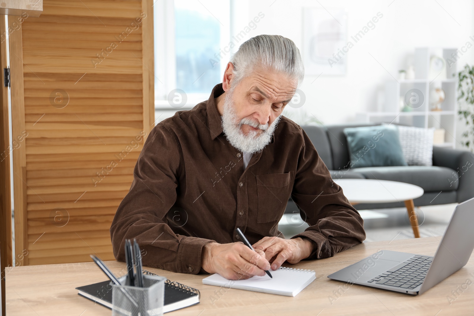 Photo of Senior man taking notes while learning online at desk indoors. Self-study