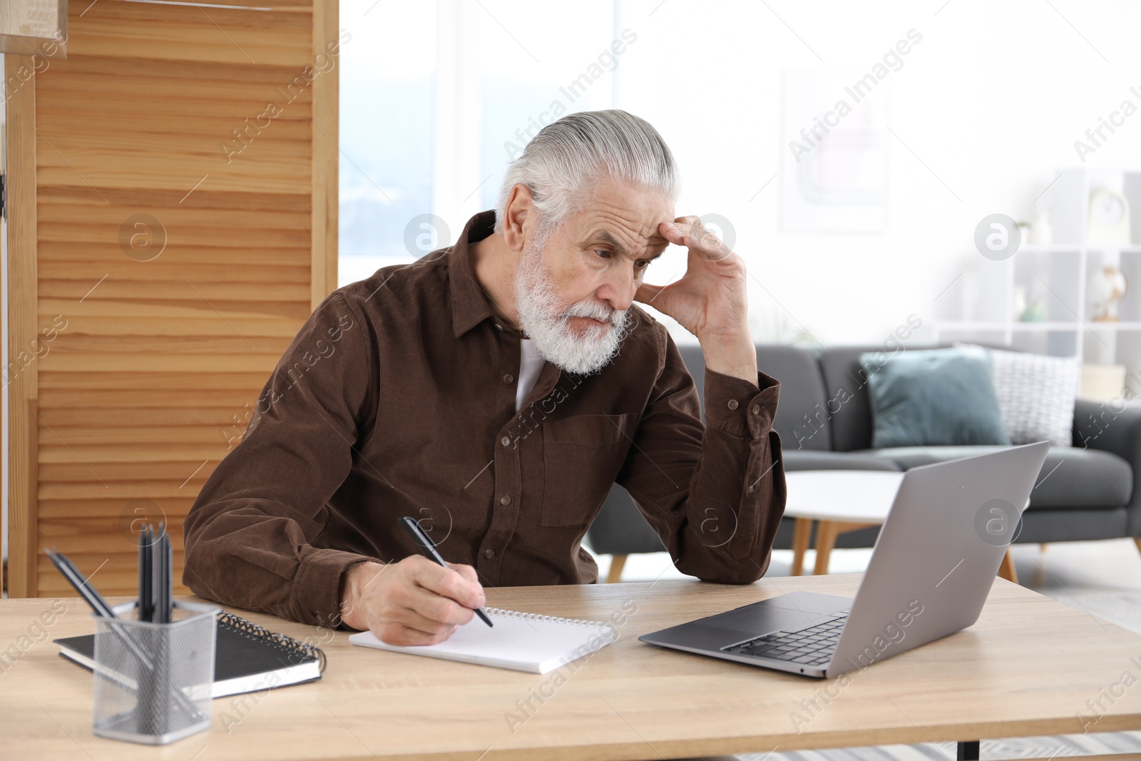 Photo of Senior man taking notes while learning online at desk indoors. Self-study