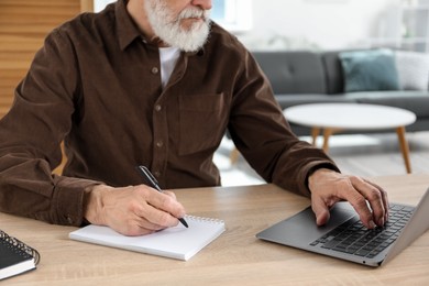 Photo of Senior man taking notes while learning online at desk indoors, closeup. Self-study