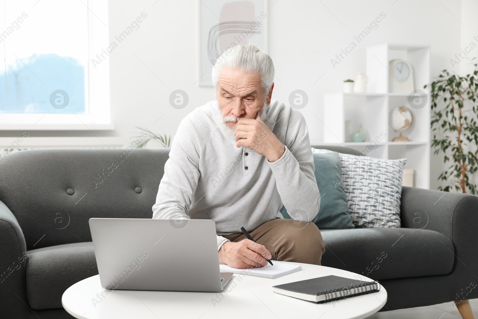 Photo of Senior man taking notes while learning online at table indoors. Self-study