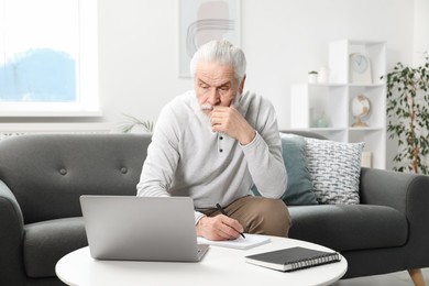 Photo of Senior man taking notes while learning online at table indoors. Self-study