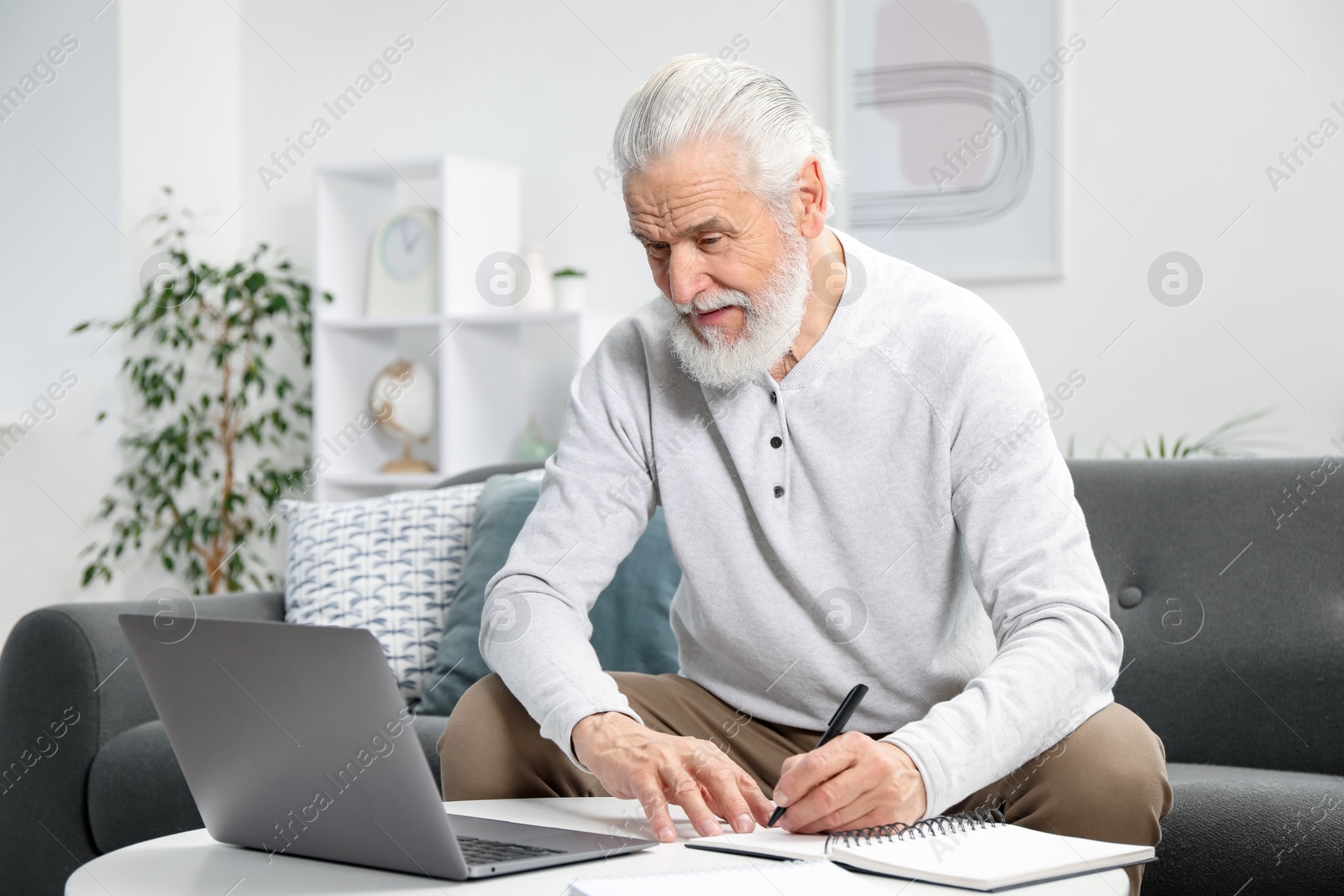 Photo of Senior man taking notes while learning online at table indoors. Self-study