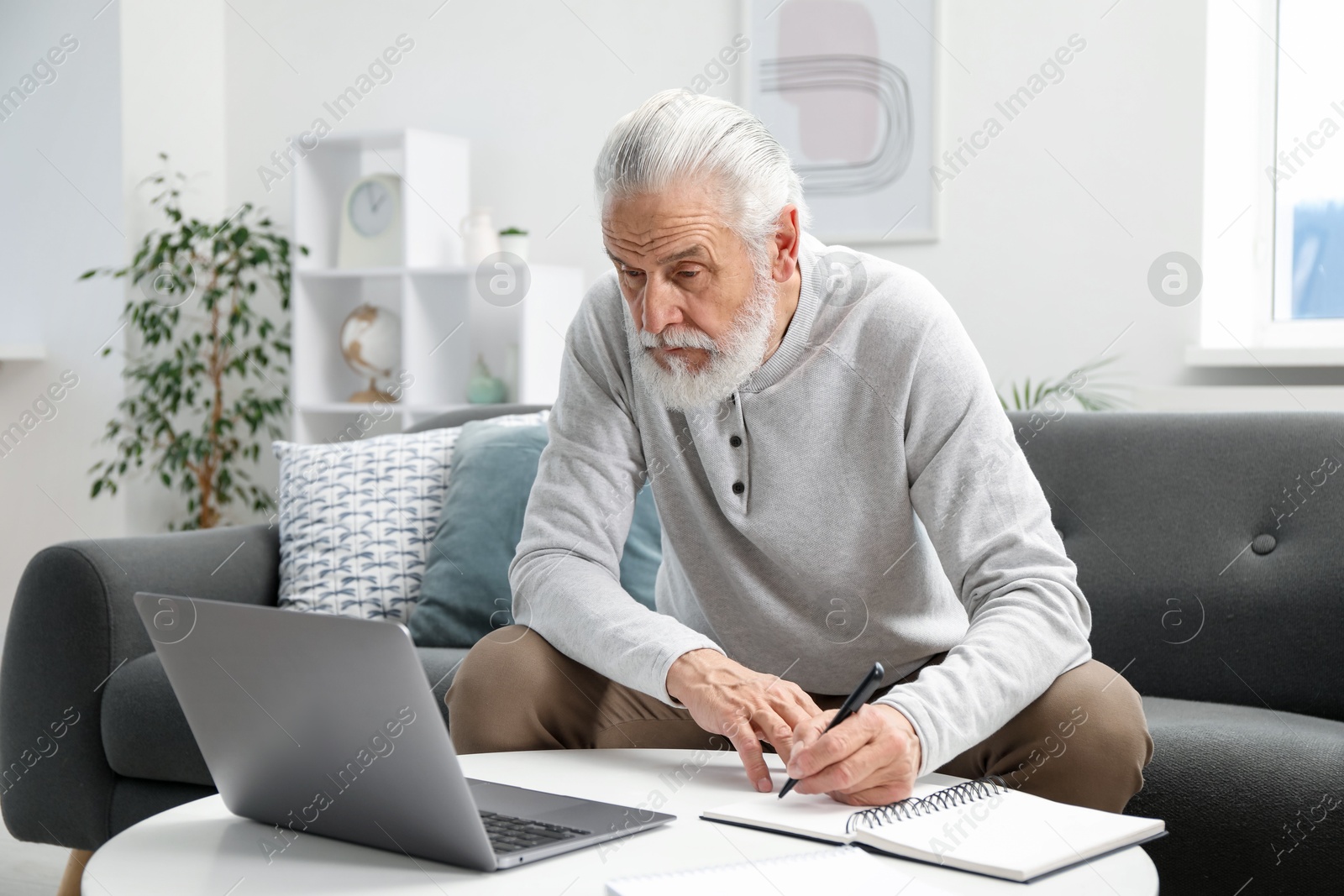 Photo of Senior man taking notes while learning online at table indoors. Self-study