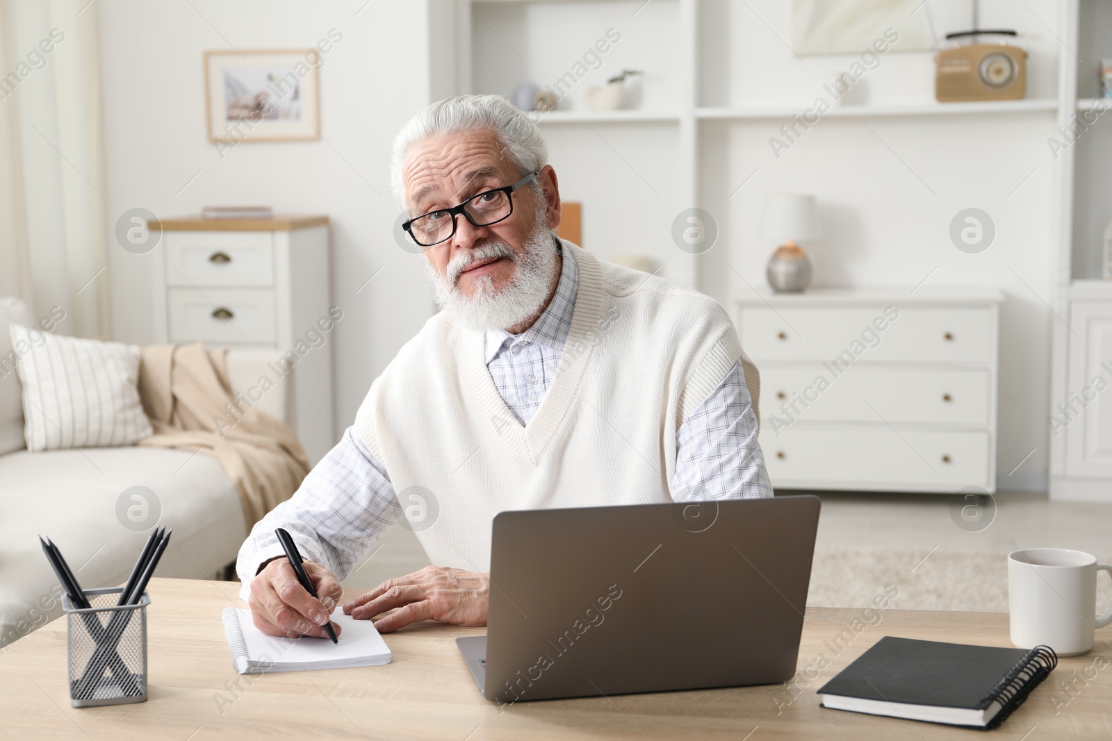 Photo of Senior man taking notes while learning online at desk indoors. Self-study