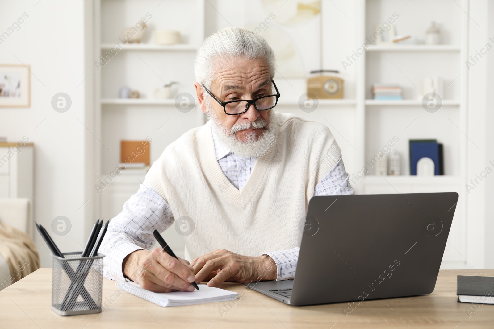 Photo of Senior man taking notes while learning online at desk indoors. Self-study