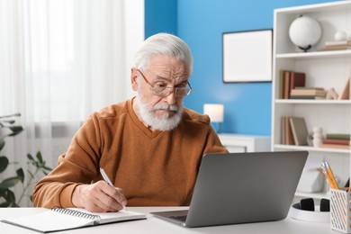 Photo of Senior man taking notes while learning online at desk indoors. Self-study