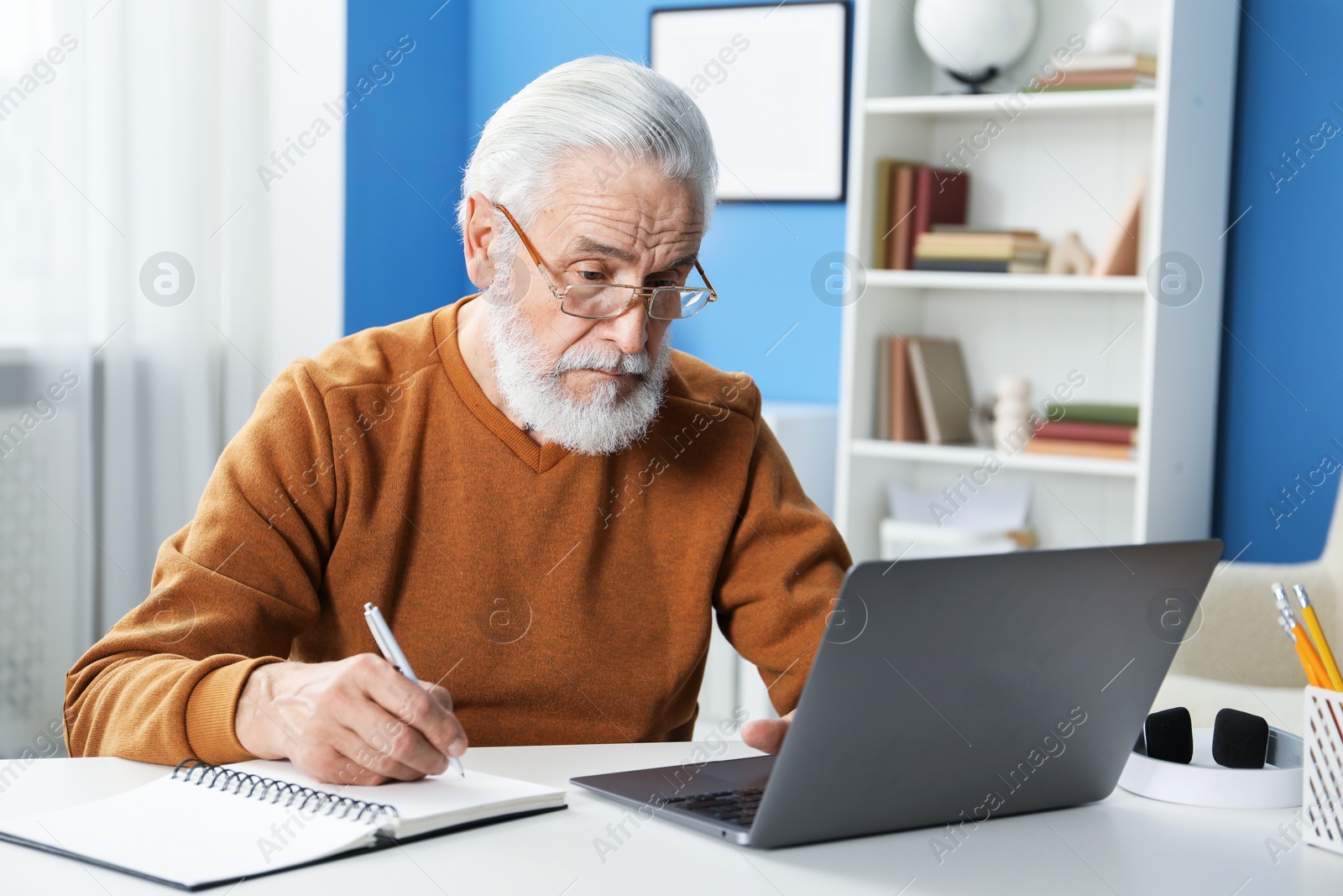 Photo of Senior man taking notes while learning online at desk indoors. Self-study