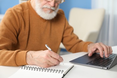 Photo of Senior man taking notes while learning online at desk indoors, closeup. Self-study