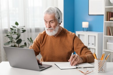 Photo of Senior man taking notes while learning online at desk indoors. Self-study