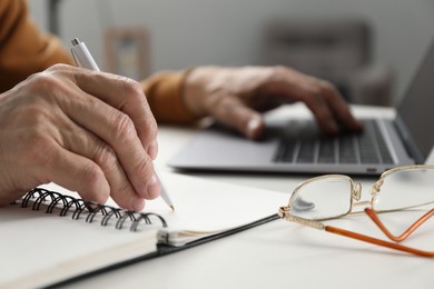 Photo of Senior man taking notes while learning online at desk indoors, closeup. Self-study