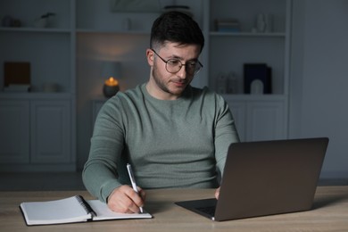 Photo of Man taking notes during online lesson at desk indoors in evening. Self-study