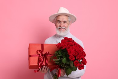 Photo of Senior man with bouquet of red roses and gift box on pink background