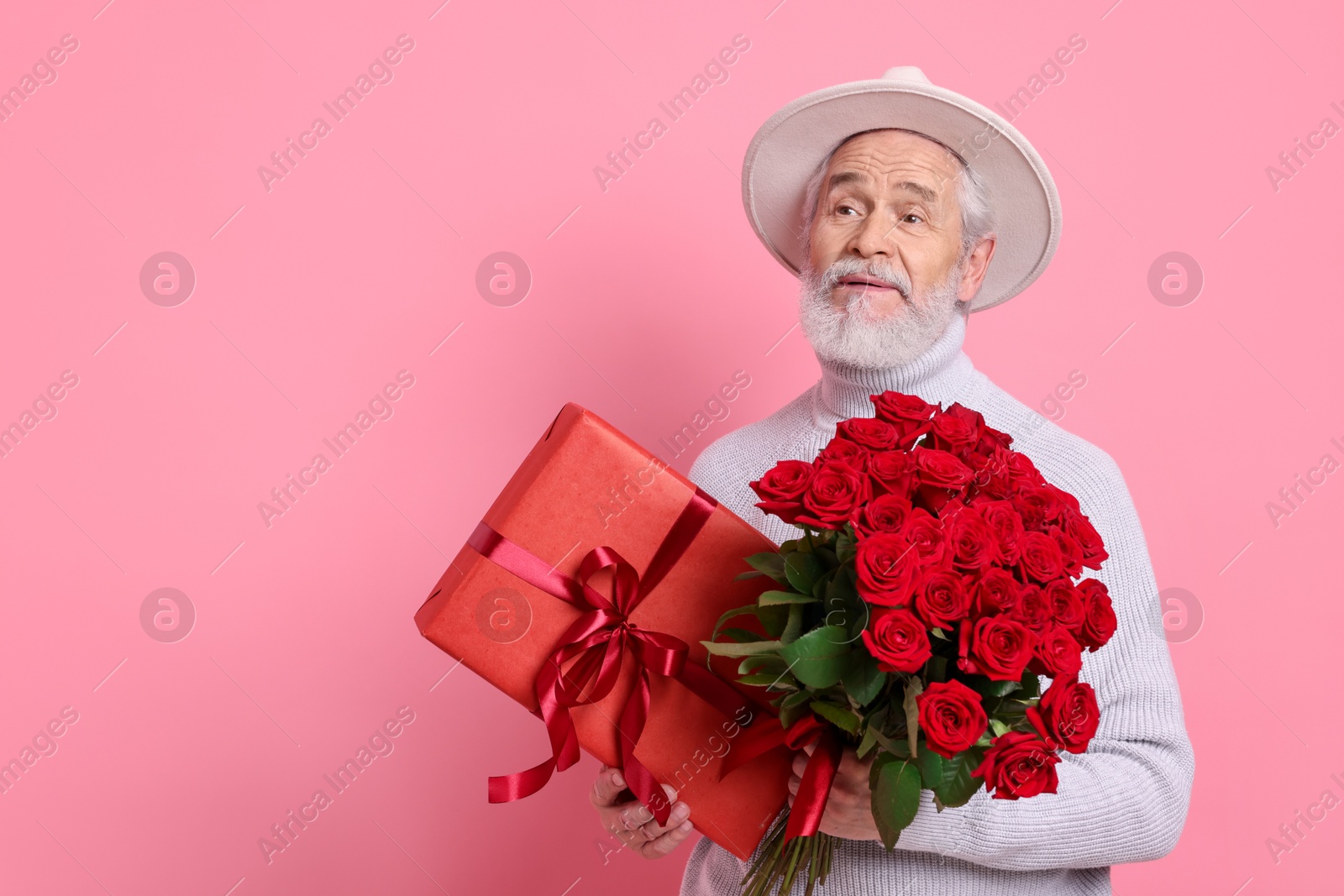 Photo of Senior man with bouquet of red roses and gift box on pink background, space for text