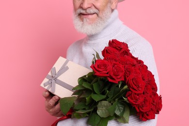 Photo of Senior man with bouquet of red roses and gift box on pink background, closeup