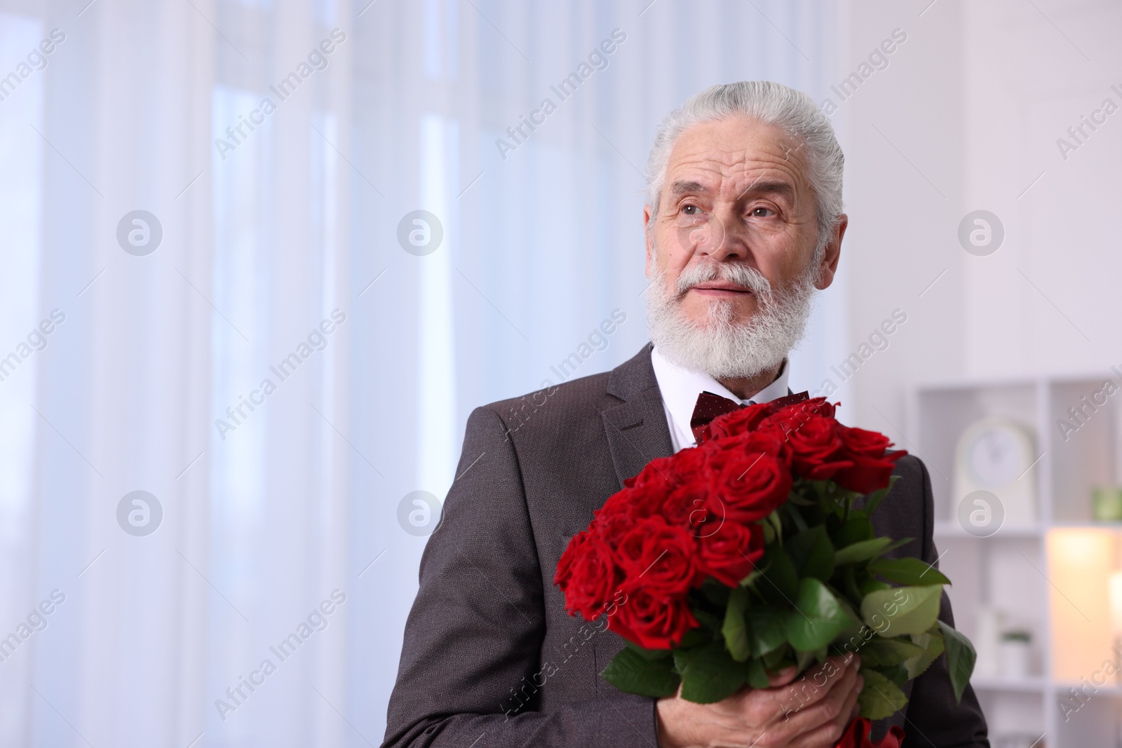 Photo of Senior man with bouquet of red roses at home, space for text