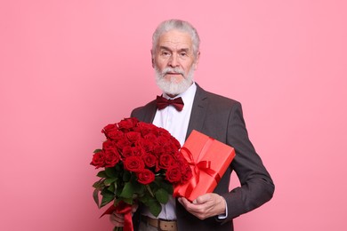 Photo of Senior man with bouquet of red roses and gift box on pink background