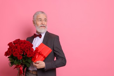 Photo of Senior man with bouquet of red roses and gift box on pink background, space for text