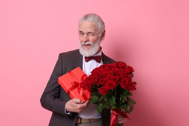 Photo of Senior man with bouquet of red roses and gift box on pink background