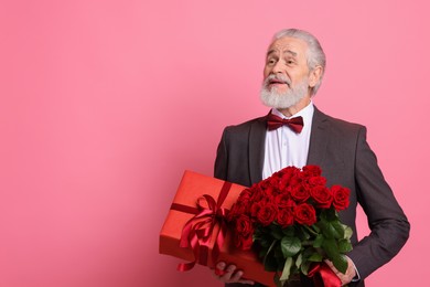 Photo of Senior man with bouquet of red roses and gift box on pink background, space for text