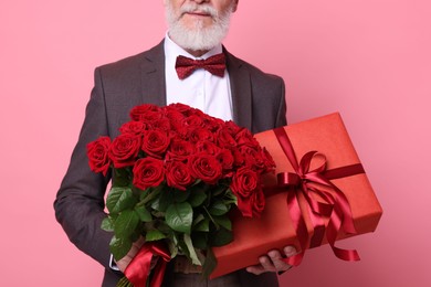 Photo of Senior man with bouquet of red roses and gift box on pink background, closeup