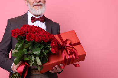 Photo of Senior man with bouquet of red roses and gift box on pink background, closeup. Space for text