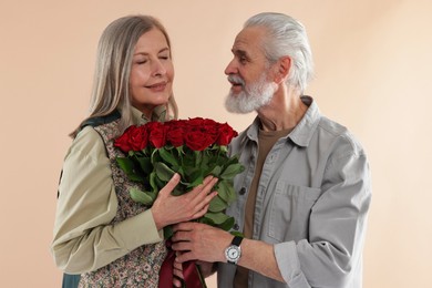 Photo of Man presenting bouquet of red roses to his happy wife on beige background