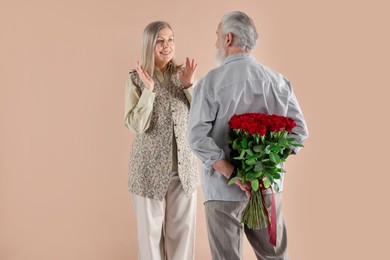 Photo of Man hiding bouquet of red roses for his happy wife on beige background, back view