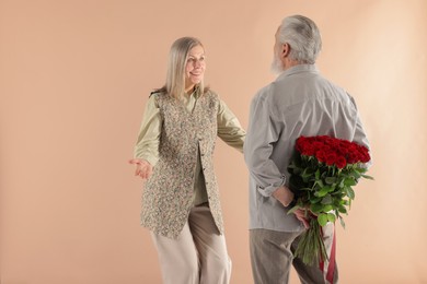 Photo of Man hiding bouquet of red roses for his happy wife on beige background, back view