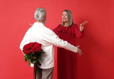 Photo of Man hiding bouquet of roses for his happy wife on red background, back view