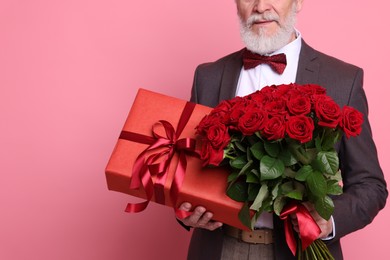 Photo of Senior man with bouquet of red roses and gift box on pink background, closeup. Space for text