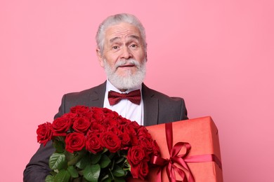 Photo of Senior man with bouquet of red roses and gift box on pink background