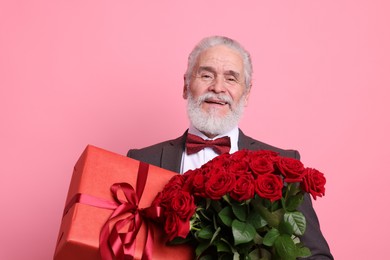 Photo of Senior man with bouquet of red roses and gift box on pink background