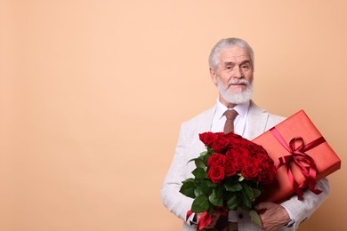 Photo of Senior man with bouquet of red roses and gift box on beige background, space for text