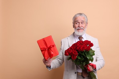 Photo of Senior man with bouquet of red roses and gift box on beige background, space for text