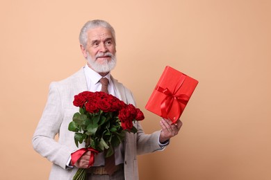 Photo of Senior man with bouquet of red roses and gift box on beige background, space for text