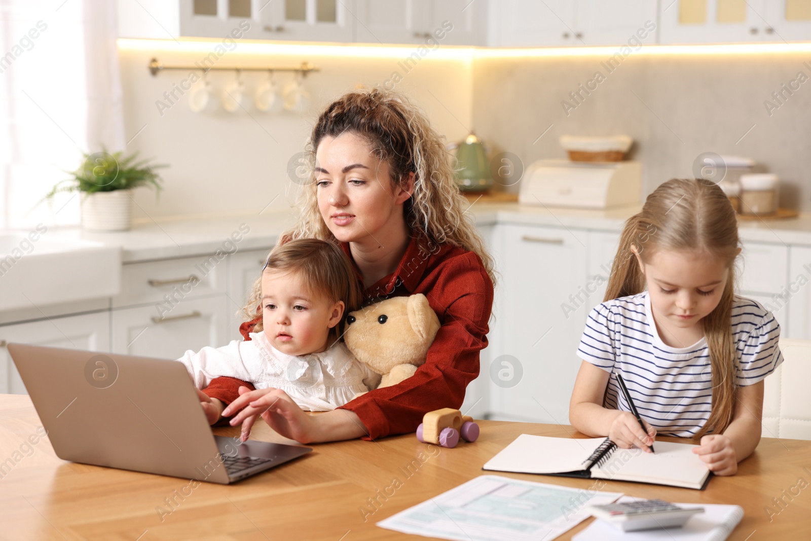 Photo of Single mother working with laptop and her daughters at table in kitchen