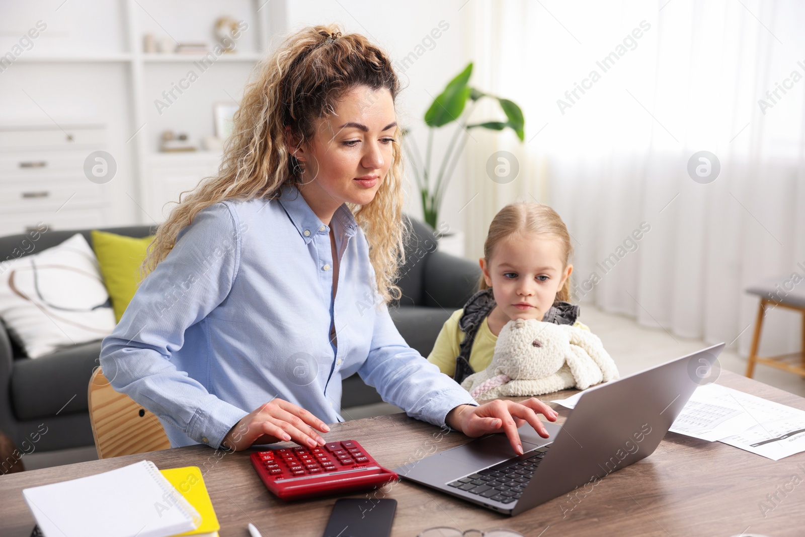 Photo of Single mother using calculator while working on laptop and her daughter at table indoors