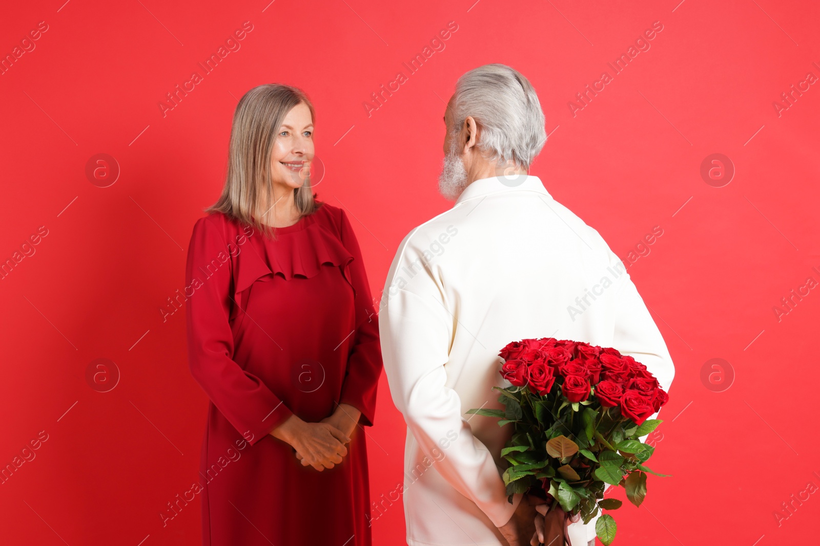 Photo of Man hiding bouquet of roses for his happy wife on red background, back view