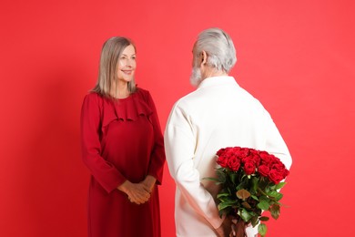 Photo of Man hiding bouquet of roses for his happy wife on red background, back view