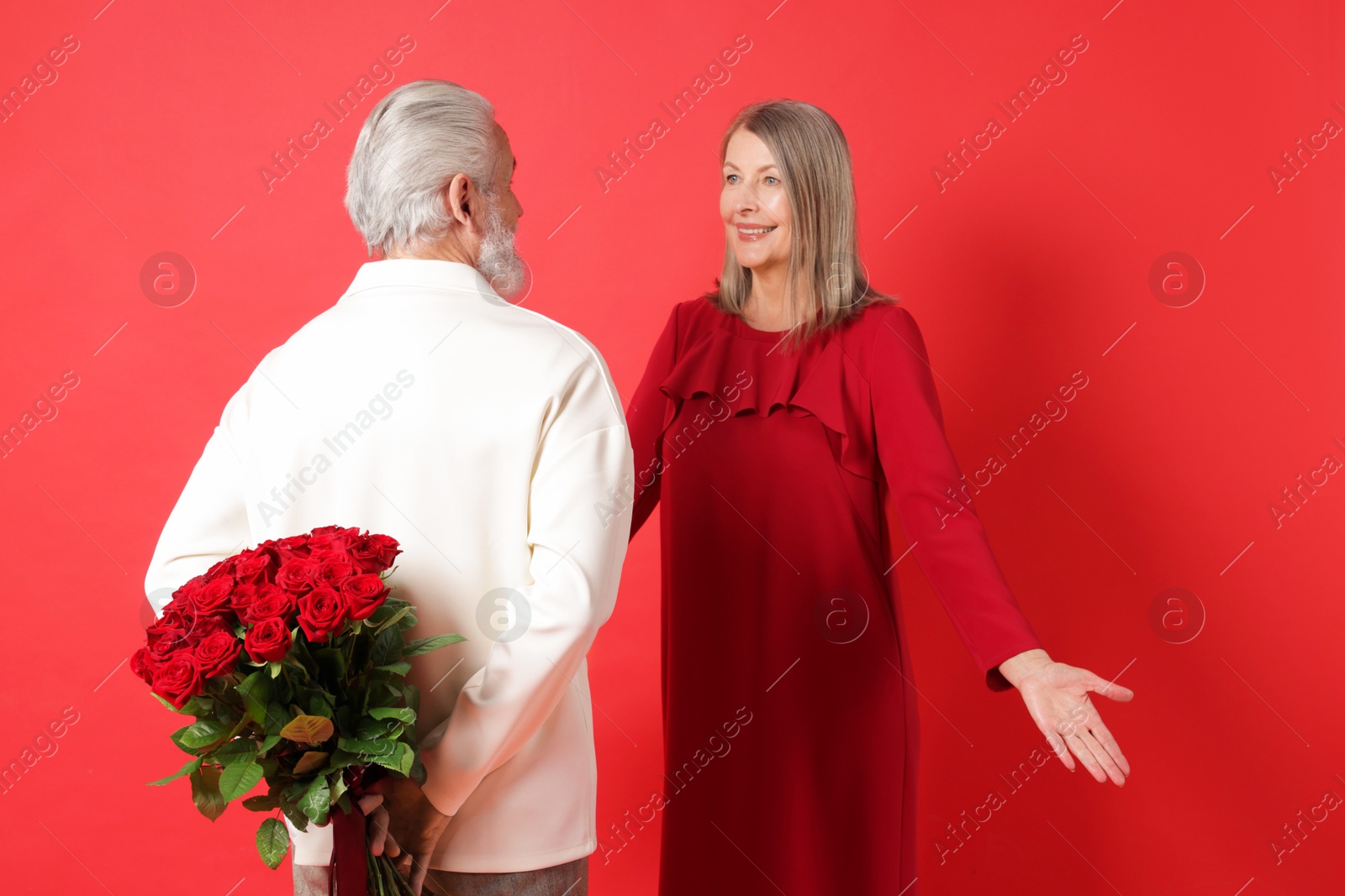 Photo of Man hiding bouquet of roses for his happy wife on red background, back view