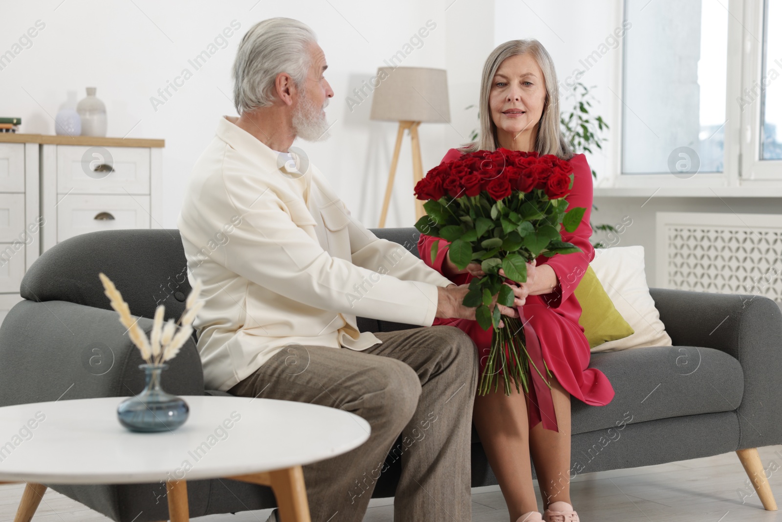 Photo of Man presenting bouquet of roses to his wife on sofa at home