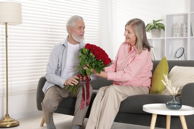 Photo of Man presenting bouquet of roses to his happy wife on sofa at home